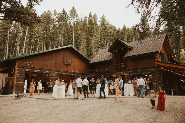A rustic wooden wedding venue building surrounded by tall pine trees with a group of wedding guests and a bride standing on the patio.