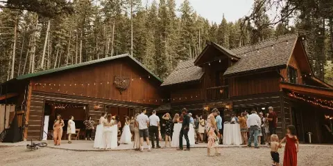 A rustic wooden wedding venue building surrounded by tall pine trees with a group of wedding guests and a bride standing on the patio.