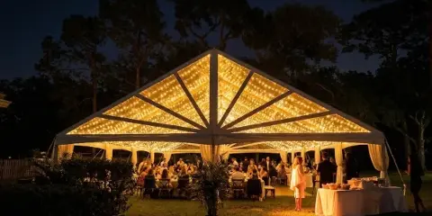 A large white event tent illuminated with string lights at night, set up for a reception with tables and guests.