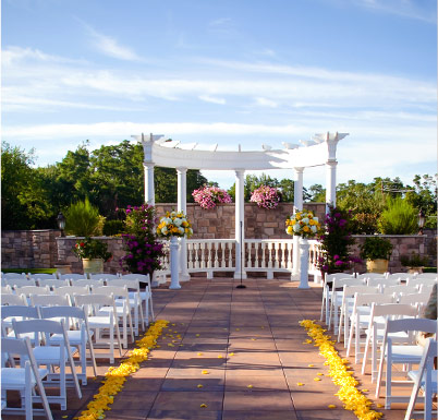 Outdoor wedding ceremony setup featuring a white pergola, floral arrangements, and rows of white chairs on a stone patio.