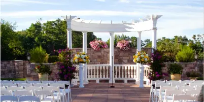 Outdoor wedding ceremony setup featuring a white pergola, floral arrangements, and rows of white chairs on a stone patio.
