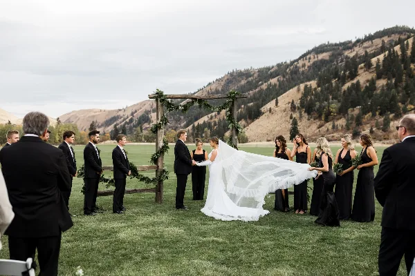 Outdoor wedding ceremony with a bride and groom at a rustic wooden arch, surrounded by a wedding party and mountain scenery.