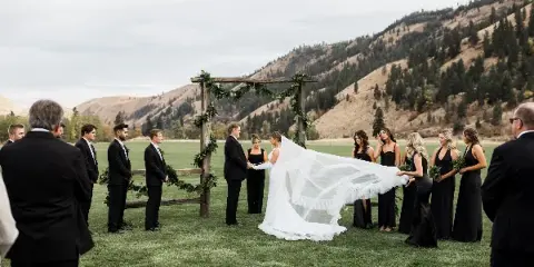 Outdoor wedding ceremony with a bride and groom at a rustic wooden arch, surrounded by a wedding party and mountain scenery.
