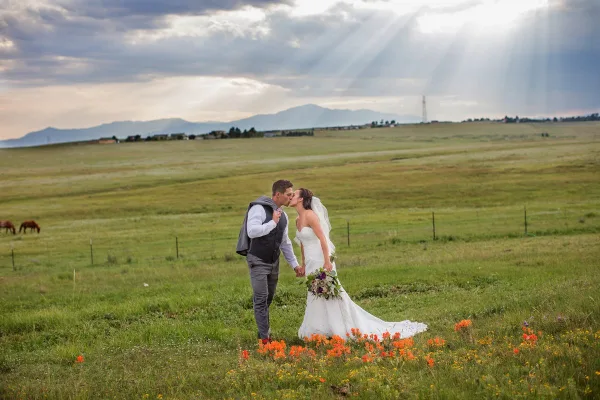 A bride and groom kissing in a grassy field with wildflowers and mountains in the background.