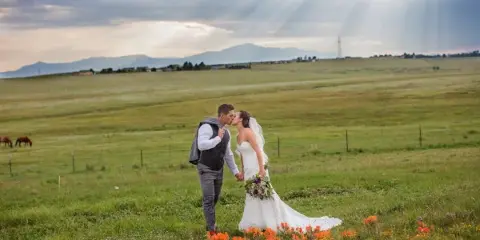 A bride and groom kissing in a grassy field with wildflowers and mountains in the background.