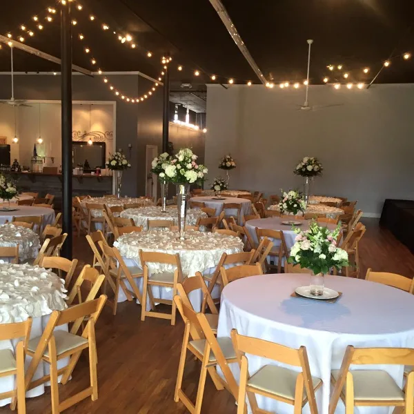 Interior of an event venue featuring round tables with white linens, floral centerpieces, and string lights hanging from the ceiling.
