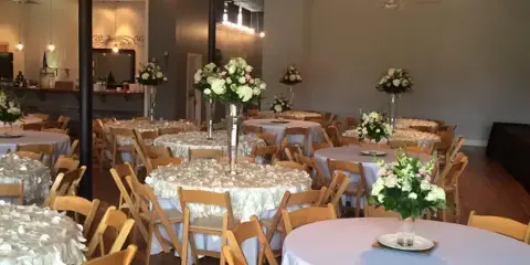 Interior of an event venue featuring round tables with white linens, floral centerpieces, and string lights hanging from the ceiling.