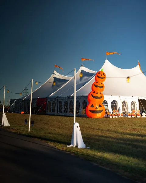 A large white event tent with arched windows and string lights set up on a grassy field, decorated with a large inflatable pumpkin stack.