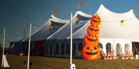A large white event tent with arched windows and string lights set up on a grassy field, decorated with a large inflatable pumpkin stack.
