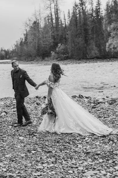A black and white photograph of a bride and groom holding hands on a rocky riverbank with a forest in the background.