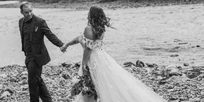 A black and white photograph of a bride and groom holding hands on a rocky riverbank with a forest in the background.