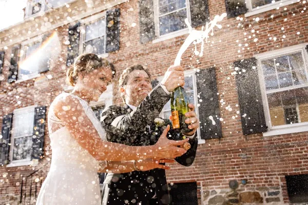 A bride and groom in wedding attire joyfully popping a bottle of champagne outdoors in front of a brick building.