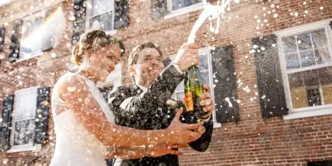 A bride and groom in wedding attire joyfully popping a bottle of champagne outdoors in front of a brick building.