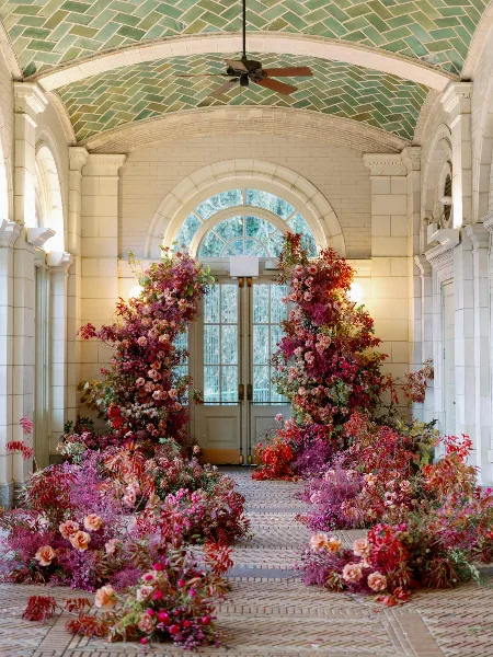 A grand floral installation featuring lush red, pink, and purple flowers arranged in arches and floor clusters within a tiled venue hallway.