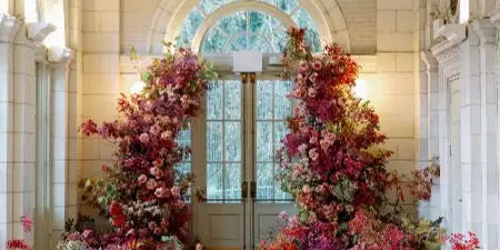 A grand floral installation featuring lush red, pink, and purple flowers arranged in arches and floor clusters within a tiled venue hallway.