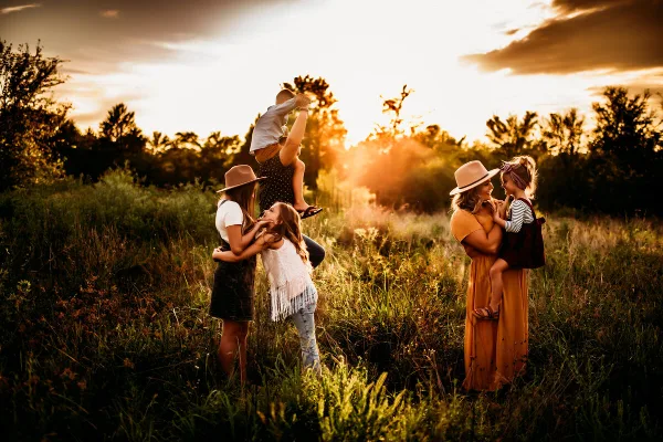 A family of five posing in a grassy field at sunset, with a mother holding a toddler and another woman holding a young child.