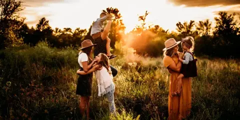 A family of five posing in a grassy field at sunset, with a mother holding a toddler and another woman holding a young child.
