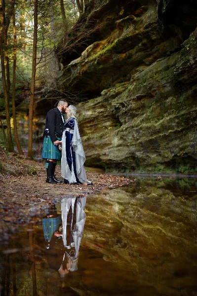 A couple in formal wedding attire sharing a kiss by a natural rock formation and water reflection.