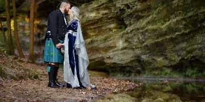 A couple in formal wedding attire sharing a kiss by a natural rock formation and water reflection.