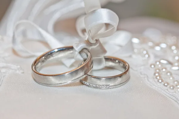 A close-up of two wedding rings resting on a white fabric with a veil and pearls in the background.