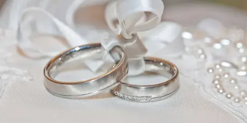A close-up of two wedding rings resting on a white fabric with a veil and pearls in the background.