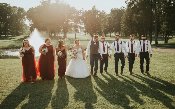 A wedding party, including the bride and groom, walking on a grassy lawn near a fountain.