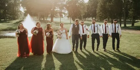 A wedding party, including the bride and groom, walking on a grassy lawn near a fountain.