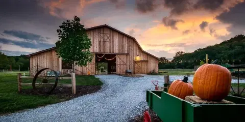 Exterior view of a rustic wooden barn wedding venue at sunset with a gravel driveway and pumpkins in the foreground.