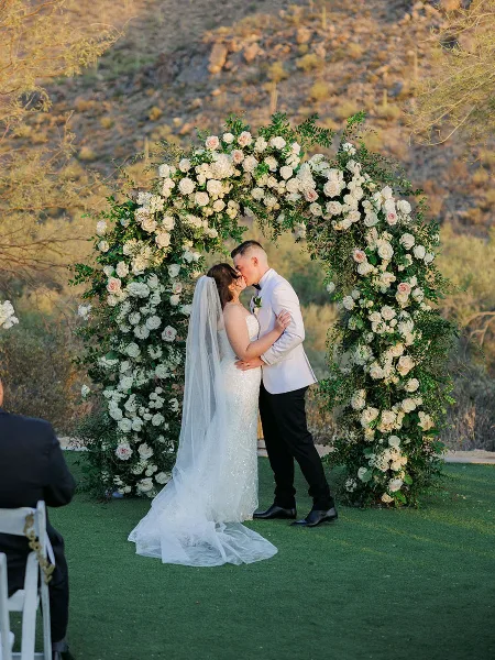 A bride and groom kissing in front of a large floral arch made of white and pink roses and greenery at an outdoor wedding venue.