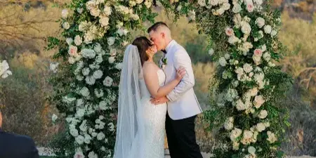 A bride and groom kissing in front of a large floral arch made of white and pink roses and greenery at an outdoor wedding venue.