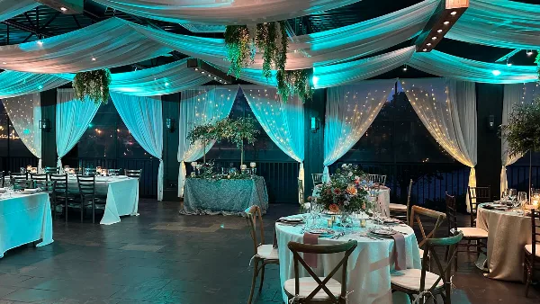 Interior view of a wedding venue featuring white ceiling drapery, string lights, and round tables set with white linens and wooden chairs.