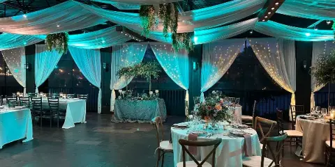 Interior view of a wedding venue featuring white ceiling drapery, string lights, and round tables set with white linens and wooden chairs.