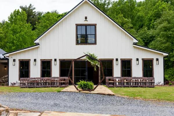 Exterior view of a white modern rustic barn with a wooden ceremony arch and rows of chairs set up on a gravel path.