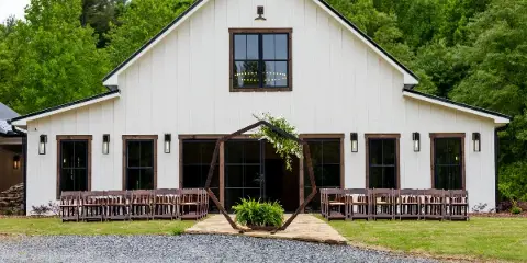 Exterior view of a white modern rustic barn with a wooden ceremony arch and rows of chairs set up on a gravel path.