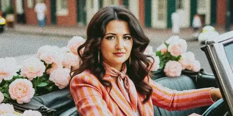 A woman in a pink striped blazer sitting in a vintage convertible car decorated with large pink floral arrangements.