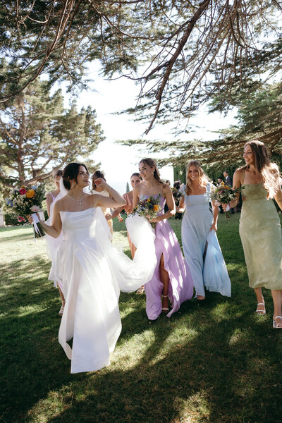 A bride in a white gown and her bridesmaids in pastel dresses walking on a grassy lawn holding bouquets.