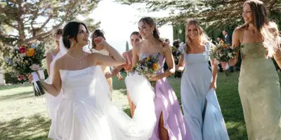 A bride in a white gown and her bridesmaids in pastel dresses walking on a grassy lawn holding bouquets.