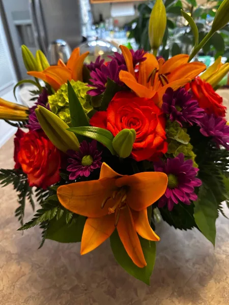 A vibrant floral arrangement featuring orange lilies, red roses, and purple chrysanthemums.