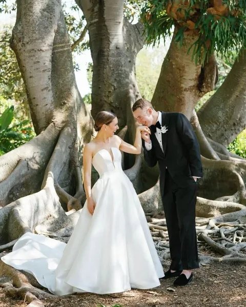 A bride in a white gown and a groom in a black tuxedo standing together outdoors near a large tree.