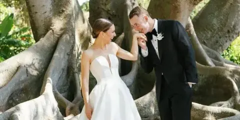 A bride in a white gown and a groom in a black tuxedo standing together outdoors near a large tree.