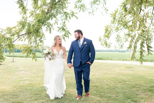 A bride and groom walking hand-in-hand on a grassy lawn under a tree.