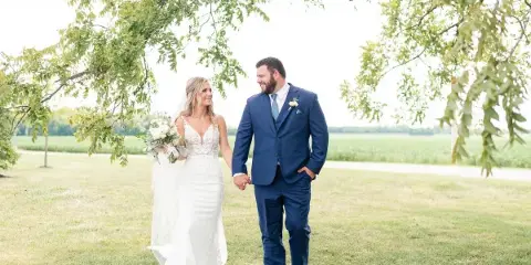 A bride and groom walking hand-in-hand on a grassy lawn under a tree.