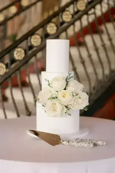 A white two-tier wedding cake decorated with white roses and greenery, displayed on a table with a cake server.