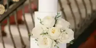 A white two-tier wedding cake decorated with white roses and greenery, displayed on a table with a cake server.