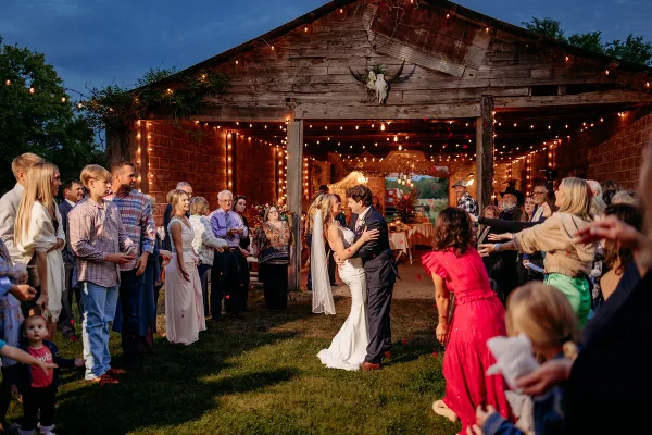 A bride and groom dancing at a rustic barn wedding reception with guests and string lights.