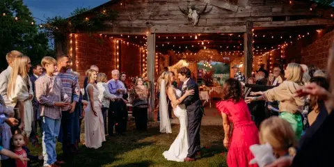 A bride and groom dancing at a rustic barn wedding reception with guests and string lights.