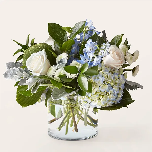 A floral arrangement featuring white roses, blue hydrangeas, and greenery in a clear glass vase.