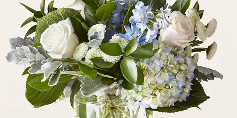 A floral arrangement featuring white roses, blue hydrangeas, and greenery in a clear glass vase.