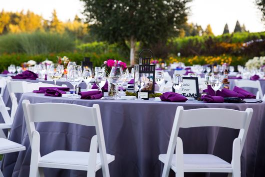 Outdoor reception table setting with white chairs, purple linens, and floral centerpieces
