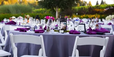 Outdoor reception table setting with white chairs, purple linens, and floral centerpieces